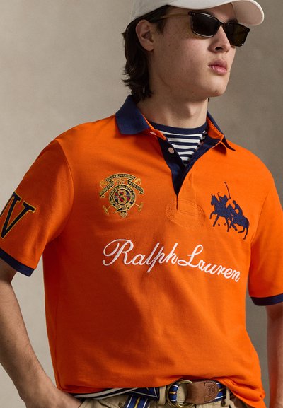 Πορτοκαλί πόλο μπλούζα με ναυτικού χρώματος λεπτομέρειες, με λογότυπο και κεντημένα αρχικά. Ριγέ πουκάμισο από μέσα, φορεμένο με ζώνη.