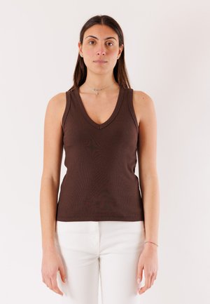 Donna con capelli lunghi e scuri, indossa una canotta marrone con scollo a V e pantaloni bianchi, in piedi davanti a uno sfondo chiaro e uniforme.