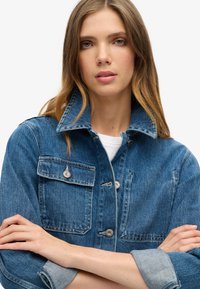 Chaqueta de mezclilla en azul medio, con un cuello alto, dos bolsillos en el pecho con cierres de metal y un corte ligeramente entallado con mangas acampanadas.