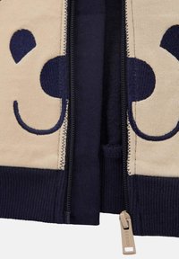 Nærbilde av beige og marineblå zip-up-stoff med broderi av et smilende ansikt og beige glidelås som er merket "GUESS."