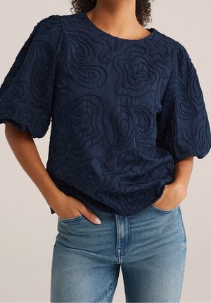 Femme portant un chemisier bleu marine texturé à manches courtes avec des motifs abstraits et un jean denim bleu clair, les mains dans les poches.