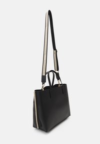 Zwarte leren tote bag met een gestructureerde vorm, voorzien van gouden hardware en een gesigneerde, logo-geprinte schouderband. Geleide zijpaneel detail.