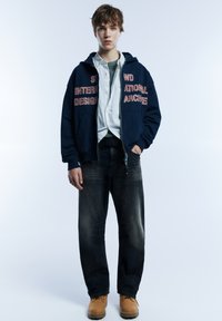 Marineblauer Kapuzenpullover mit rotem Text, weißem Hemd darunter, dunkle Jeans und beige Stiefel. Einfaches Design, lockere Passform und kontrastreiche Farben.