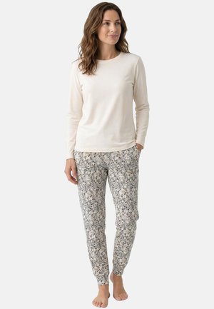 Vrouw met golvend bruin haar, gekleed in een beige top met lange mouwen en een broek met bloemmotief, staat blootsvoets met één hand in de zak.