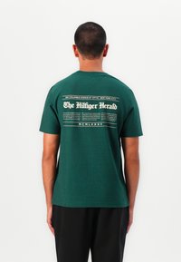 Grøn kortærmet t-shirt lavet af bomuld. Har et hvidt grafisk print på ryggen, der ligner en avisoverskrift og tekstlayout.