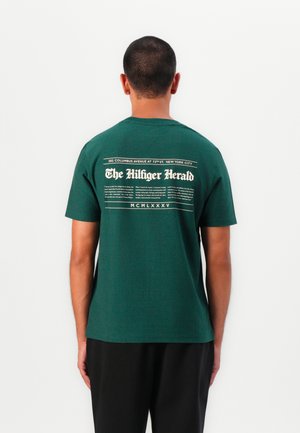 T-shirt verde a maniche corte in cotone. Presenta una stampa grafica bianca sul retro, che ricorda un titolo di giornale e una disposizione del testo.