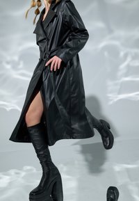 Černý trenčkot z umělé kůže s páskem v pase, širokými klopami a postranním rozparkem, doplněný černými vysokými botami na podpatku až ke kolenům.