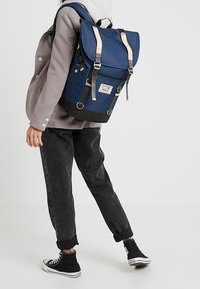 Sac à dos en tissu bleu avec détails en cuir, bretelles réglables et fermeture à enroulement. Fond noir, poche avant avec écusson logo et ferrures métalliques.