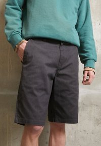 Ciemnoszare szorty o prostym kroju, wykonane z mocnego materiału. Posiadają boczne kieszenie oraz widoczne detale szwów. Model nosi tealową bluzę.
