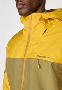 Gelbe und olivgrüne wasserdichte Outdoor-Jacke mit durchgehendem Reißverschluss, verstellbarem Kapuzenzug und "ENDURA"-Logo auf der Brust.