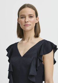 Marineblå klänning med v-ringning och lager av volangärmar. Tyget verkar slätt med en subtil lyster, vilket framhäver rena linjer och en strukturerad design.