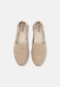 Svijetlosmeđi slip-on cipele s teksturiranom površinom, s V-oblikovanim šavom. Unutrašnjost uključuje otisnuti uzorak i logo.