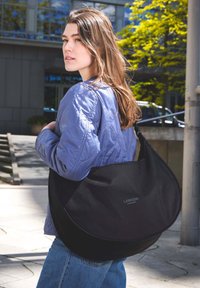 Mladá žena se světle hnědými vlasy nosí levandulovou bundu a nese velkou černou kabelku Larkson Hamburg venku ve dne.