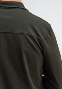 Dunkelgrünes Hemd mit Kragen, glatter Textur, sichtbaren Nahtdetails und einem eingesetzten Schulterdesign, nur von hinten sichtbar.