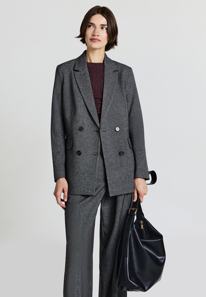 Blazer grigio doppiopetto con tessuto testurizzato e bottoni neri, abbinato a pantaloni grigi. Una borsa a tracolla nera in pelle è tenuta in una mano.