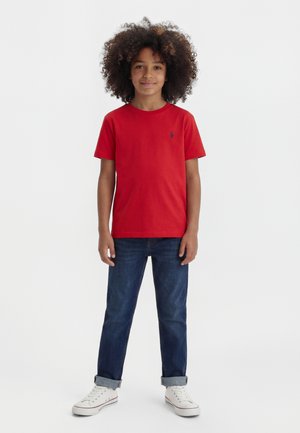 Garçon aux cheveux bouclés portant un t-shirt rouge, un jean bleu retroussé et des baskets blanches, debout devant un fond blanc.