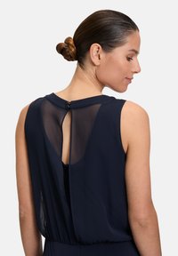 Laivastonsininen ilmanhiha Dress, jossa on läpinäkyvä takapaneeli, pyöreä kaula-aukko ja nappikiinnitys takana. Sujuva kangas ja istuva vyötärö.