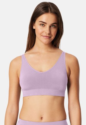 Młoda kobieta o prostych brązowych włosach, ubrana w liliowy bezrękawnik typu crop top, zwrócona przodem z delikatnym uśmiechem i neutralnym tłem.