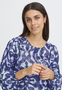 Jeune femme aux cheveux bruns, portant de grandes boucles d'oreilles en or et un chemisier à motifs bleus, serrant doucement ses mains.