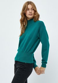 Maglione teal con collo alto, vita a coste e maniche lunghe. Realizzato in un tessuto morbido e dal design aderente. Abbinato a jeans scuri.