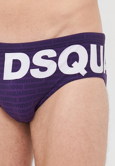 Gros plan sur la taille d'une personne portant un slip violet avec de grandes lettres blanches "DSQUA" et de plus petits motifs répétés de la marque.