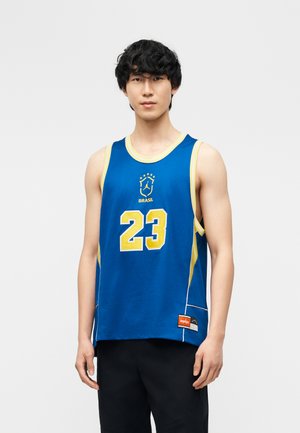 Jeune homme portant un maillot de basketball bleu et jaune du Brésil avec le numéro 23, debout devant un fond clair uni.