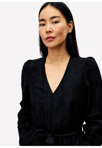 Frau mit langen dunklen Haaren, die ein schwarzes V-Ausschnitt-Kleid mit Puffärmeln und einem gebundenen Gürtel trägt, steht vor einem schlichten weißen Hintergrund.