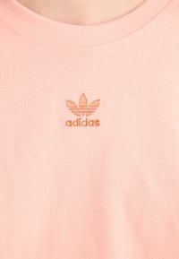 Rosa Baumwoll-T-Shirt mit Rundhalsausschnitt; verfügt über ein geprägtes orangefarbenes adidas-Logo auf der linken Seite, einfaches Design, gleichmäßige Textur.