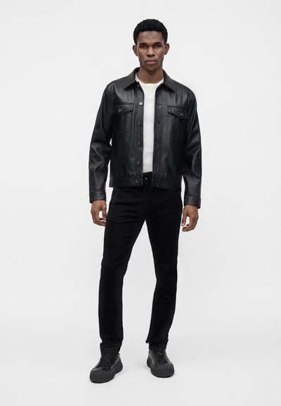 Jeune homme debout portant une veste en cuir noire, un t-shirt blanc, un pantalon noir et des baskets noires sur un fond clair uni.