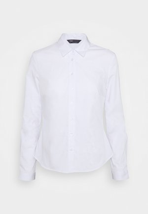 Chemise blanche à manches longues avec col pointu, boutons apparents sur le devant et coupe ajustée, présentée sur un fond uni.