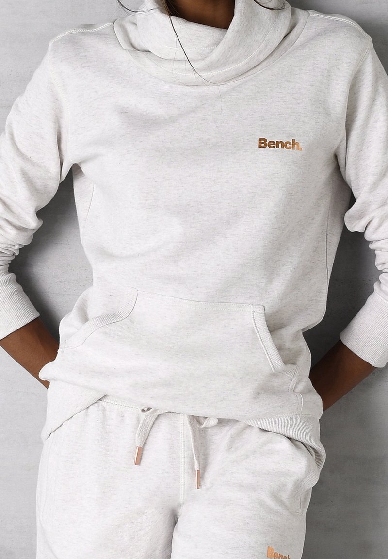 Personne portant un sweat à capuche gris clair Bench et un pantalon de jogging assorti avec des poches avant et un logo sur la poitrine et la cuisse, les mains posées sur les hanches.
