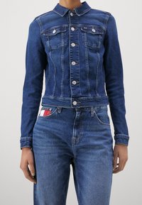 Modrá džínová bunda s límcem, předními kapsami a kovovými knoflíky. Bunda má přiléhavý střih s viditelným prošíváním a zkrácený rozměr.