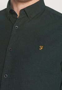 Camisa de color verde oscuro con cuello y diseño de botones, con botones negros y un pequeño logo bordado en dorado en el pecho izquierdo.