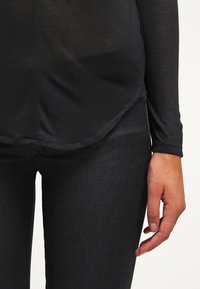 Pravá ruka uvolněná po boku, oblečená v černém topu s dlouhým rukávem a černých kalhotách na bílém pozadí.