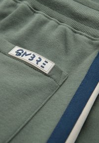 Gros plan sur un tissu vert avec une poche comportant une étiquette blanche et bleue et un détail rayé bleu et blanc sur le côté.