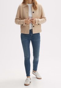 Béžová texturovaná bunda s límcem a třemi knoflíky, kombinovaná s modrými džínami a bílými teniskami. Pod ní jednoduché bílé tričko.