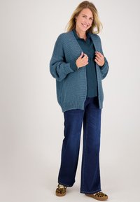 Chunky, teal gebreide cardigan met lange mouwen en een relaxte fit, gedragen over een donkerblauw button-up shirt en gecombineerd met flare jeans.