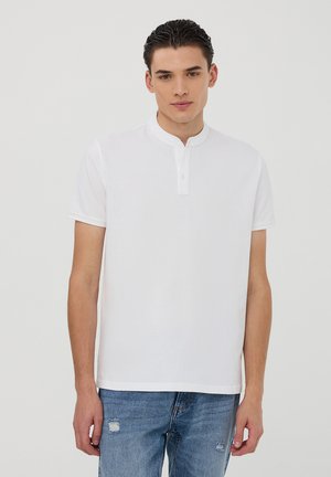 Polo shirt bianco in cotone con colletto alla cinese e una breve patta con bottoni. Indossato con jeans chiari strappati.