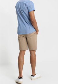 Mężczyzna w jasno-niebieskiej koszuli z krótkim rękawem, beżowych szortach do kolan i białych trampkach stoi z rękami w kieszeniach, zwrócony tyłem.