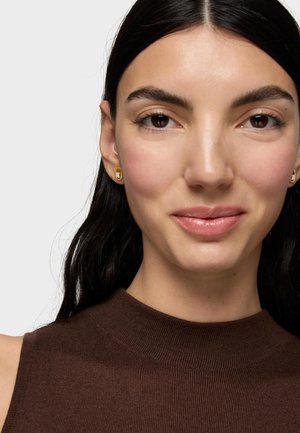 Donna con capelli scuri indossa una canotta marrone e piccoli orecchini d'oro, rivolta in avanti con un'espressione neutra su uno sfondo semplice.