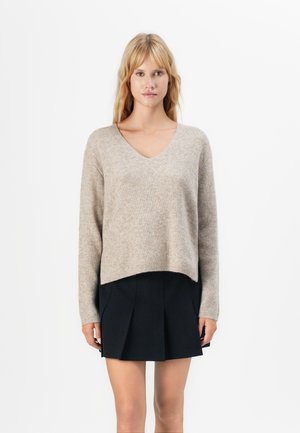 Vrouw staat gekleed in een beige geribde V-hals trui en een zwarte geplooide minirok tegen een witte achtergrond.