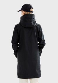 Parka noire imperméable avec capuche, coupe longue et tissu texturé. Dispose de poignets ajustables et d'un design minimaliste.