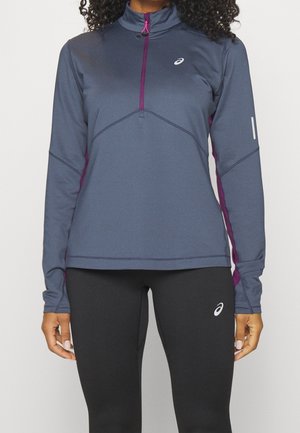 Žena nosící sportovní tričko s dlouhým rukávem v šedé a fialové barvě se čtvrtinovým zipem a černé legíny, obě s logem Asics.