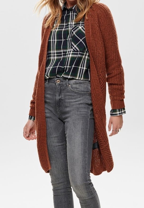 Larga chaqueta de punto color óxido con textura acanalada, de frente abierto y bolsillos laterales, superpuesta sobre una camisa a cuadros verde y negra y unos jeans grises.