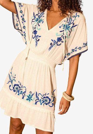 Femme portant une courte robe crème avec une broderie florale bleue, un col en V, des manches larges et une taille froncée, accessoirisée de bracelets beige épais.
