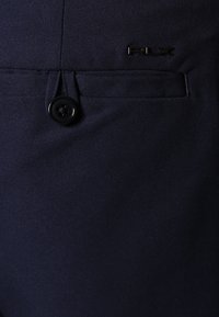 Tmavě námořnické kalhoty z hladké tkaniny, s kulatým černým knoflíkem a decentním detailem loga. Tailored kapsy s čistými okraji.