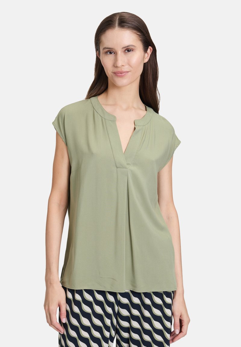 Hellgrüne Bluse mit kurzen Ärmeln, V-Ausschnitt, gefältelter Vorderseite und glatter Textur. Kombiniert mit gemusterten, weit geschnittenen Hosen in Marineblau und Creme.