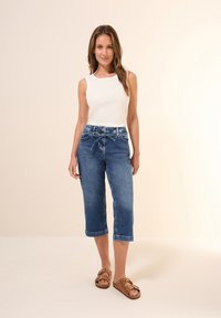 Valkoinen hihaton paita yhdistettynä puolikorkeisiin, tummansinihin denim-caprihousuihin, joissa on nyörivyö ja käänteet; ruskeat sandaalit, joissa on pilkkuja.