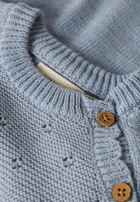 Gaiši zils adīts kardigans ar ribotu apaļu apkakli, koka pogām un nelielām rakstainām detaļām audumā. Mīksta tekstūra.