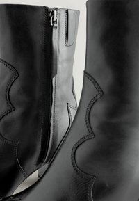 Botas de tobillo de cuero negro con un diseño ondulado cosido, que cuentan con una cremallera lateral y una textura suave. El tacón es bajo y cuadrado.